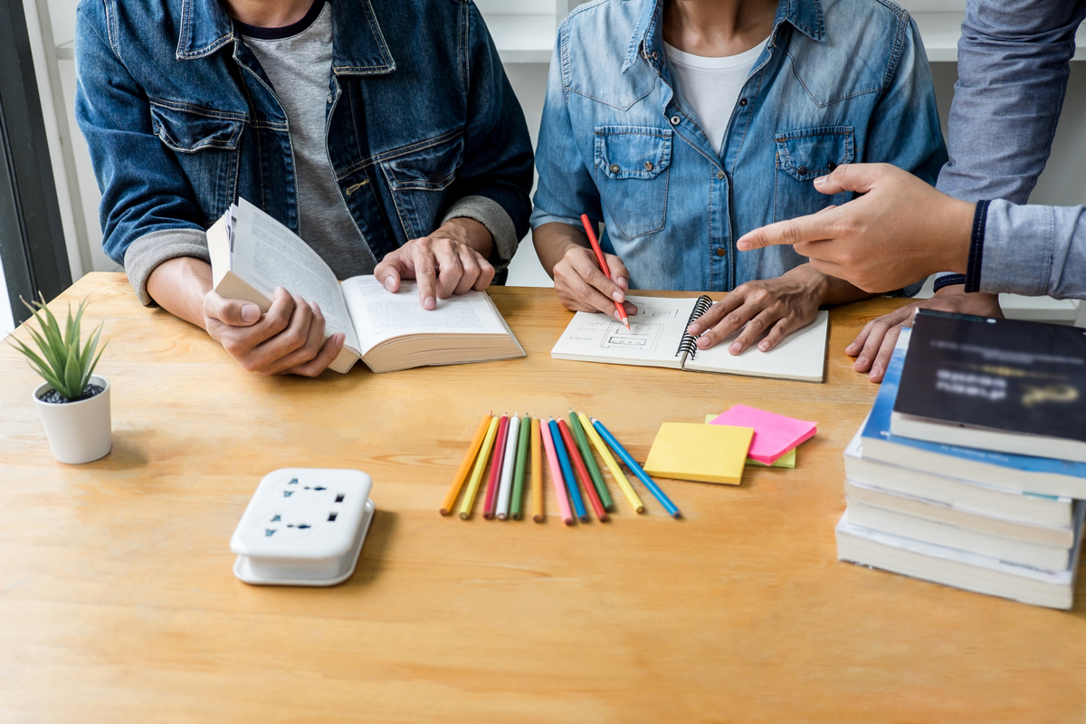 Gruppe von Gymnasiasten oder Studenten sitzt am Schreibtisch in der Bibliothek und lernt und liest, macht Hausaufgaben und übt für den Unterricht