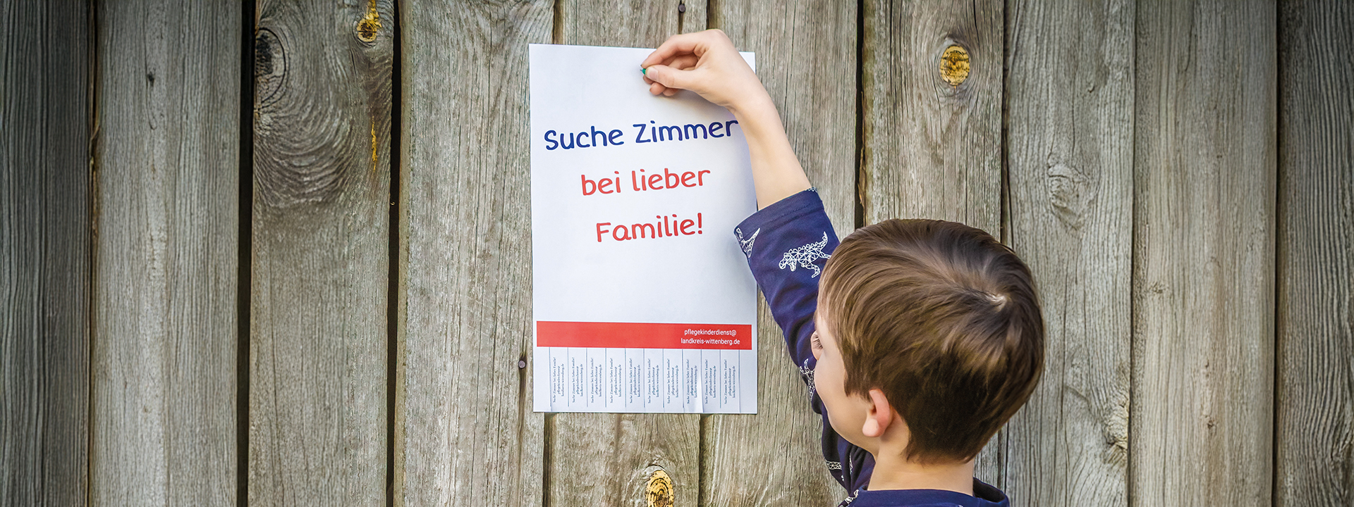 Kind pint Zettel mit der Überschrift Suche Zimmer bei lieber Familie an einer Holzwand,