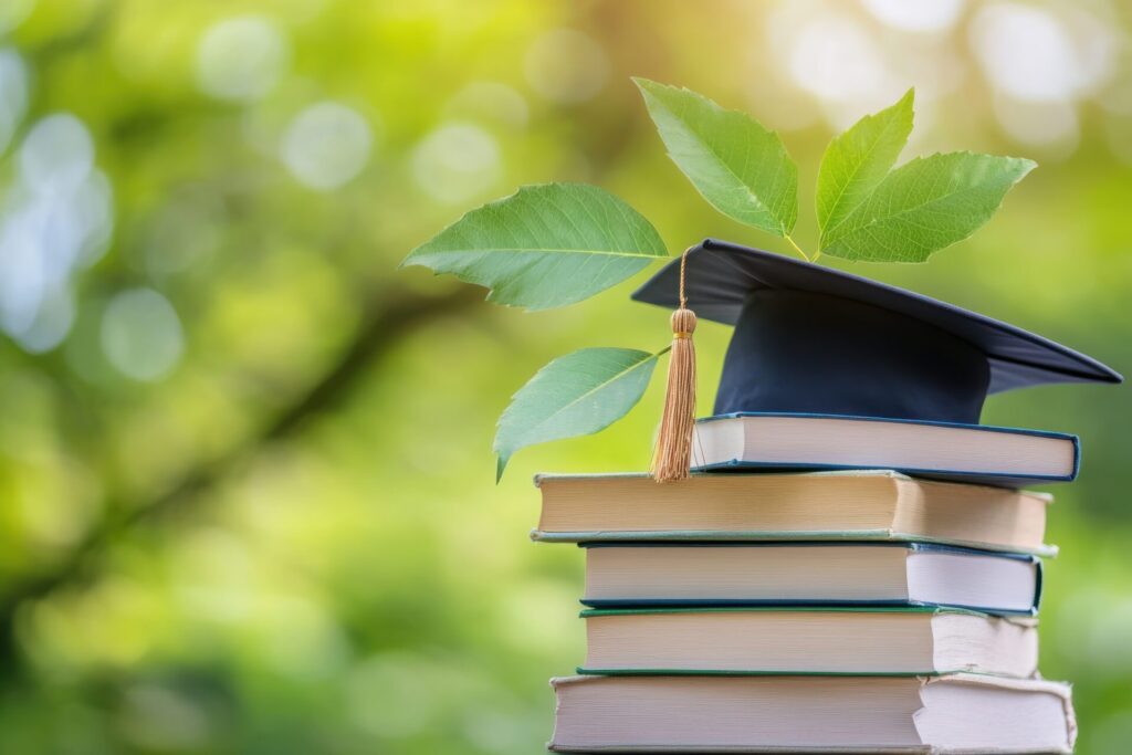 Bücherstapel mit Absolventenhut im Vordergrund rechts, im Hintergrund unscharf grün von Bäumen