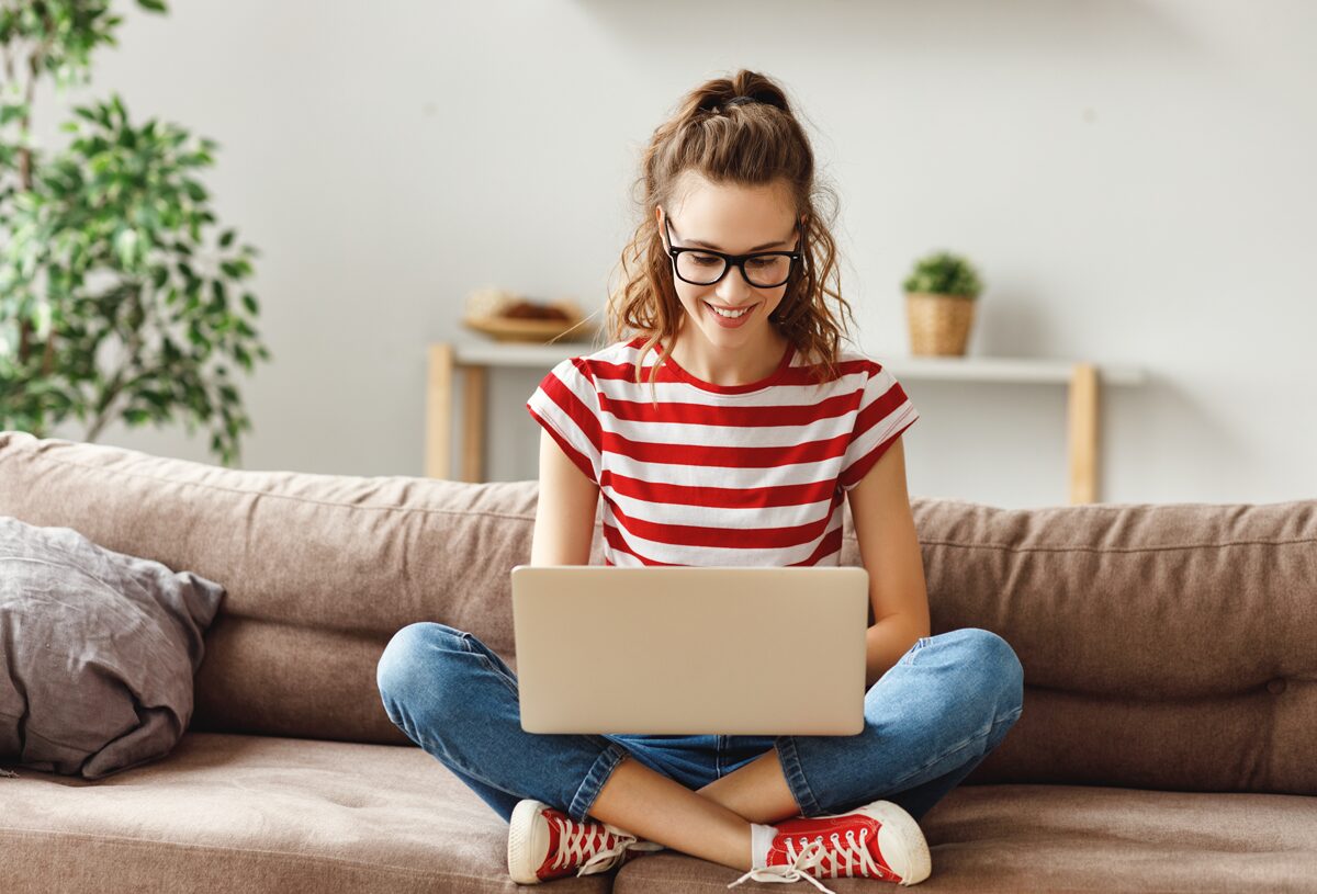 junge Frau sitzt auf einem Sofa im Schneidersitz und schaut auf ihren Laptop