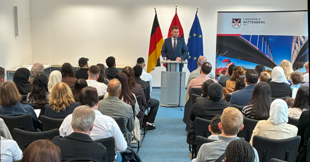 Eine Gruppe Menschen sitzt im Saal, sie sind von hinten zu sehen. in der Mitte steht vorn Landrat Christian Tylsch.