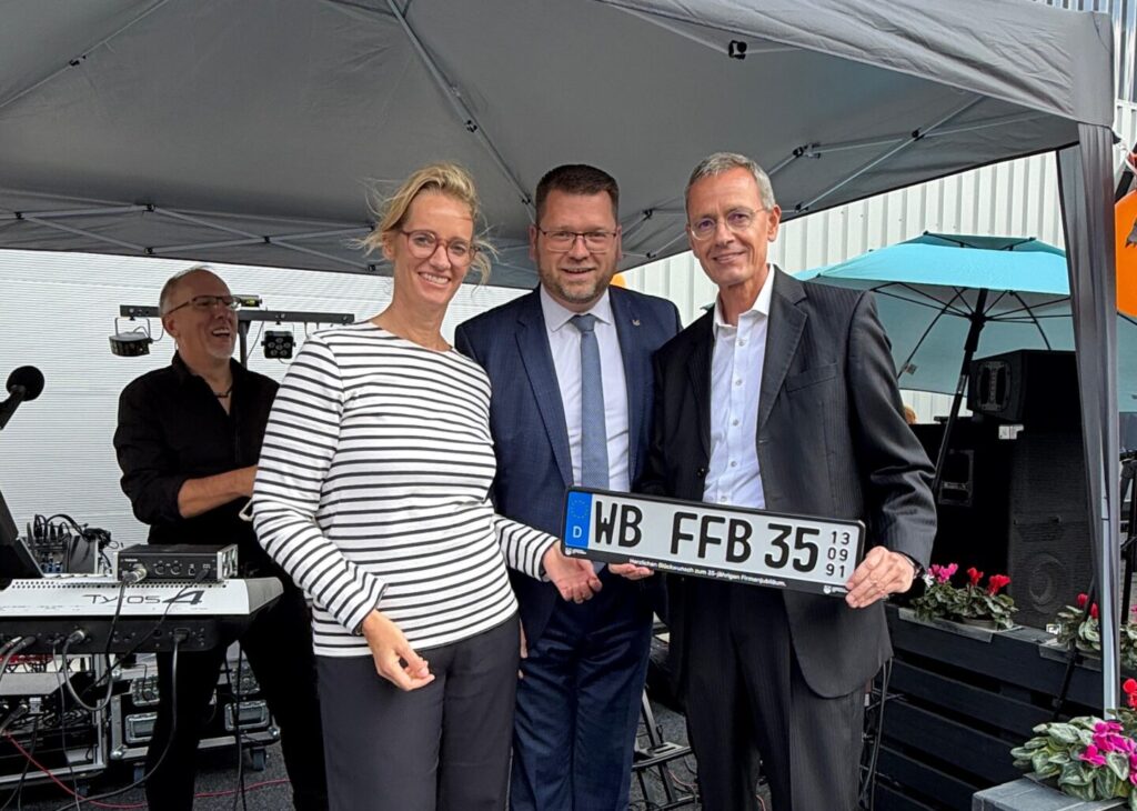 Drei Menschen halten ein Jubiläums-Nummernschild in den Händen.
