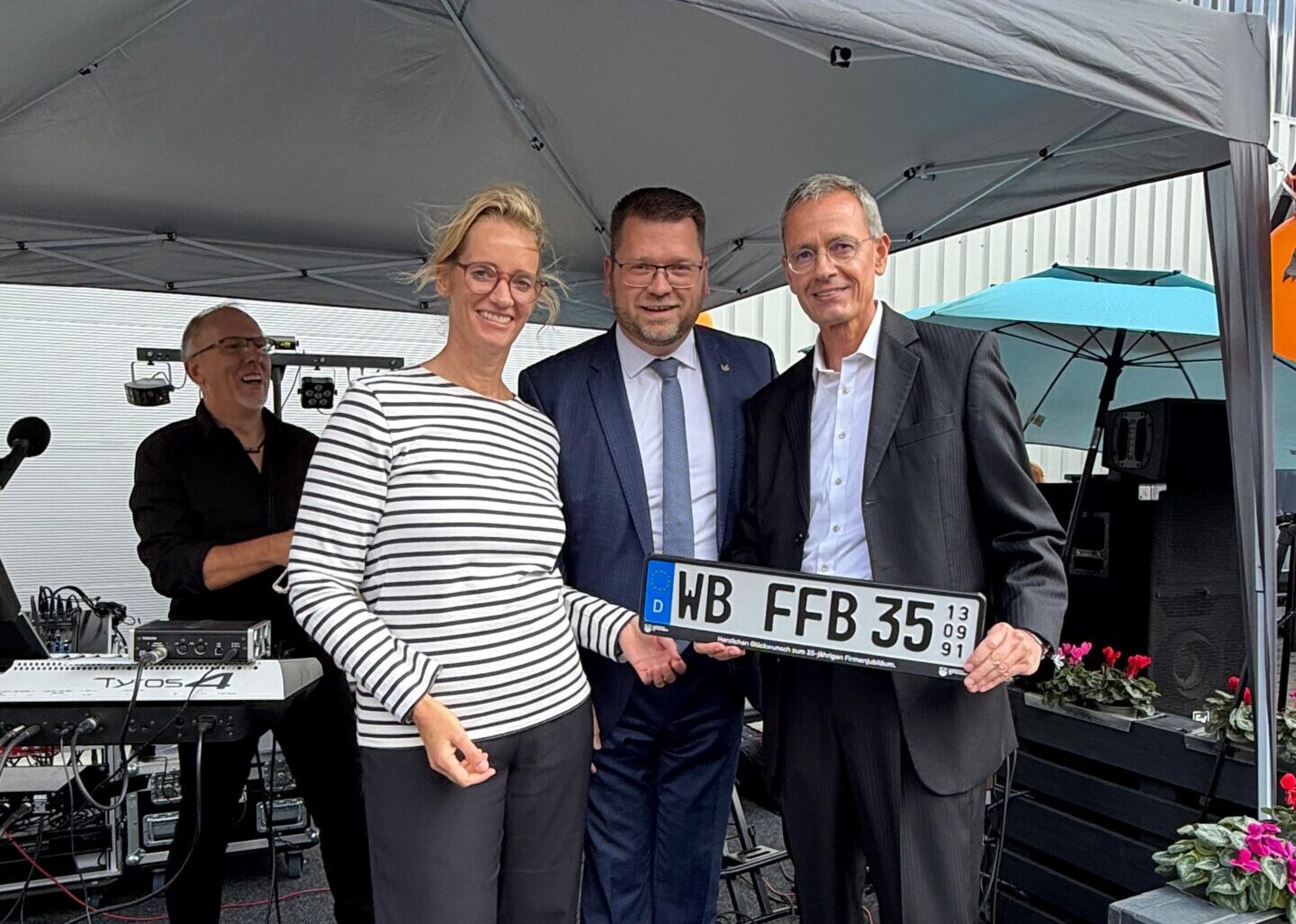 Drei Menschen halten ein Jubiläums-Nummernschild in den Händen.