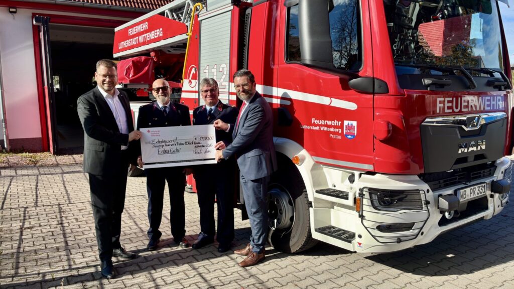 Vier Männer stehen vor einem Feuerwehrauto und halten einen Scheck hoch.