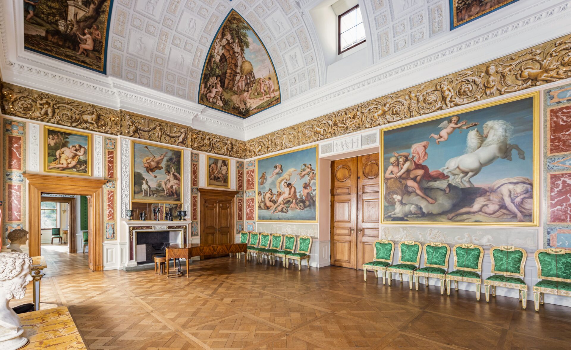 Prunkvoller Festsaal mit bemalten Decken- und Wandgemälden, grünen Stühlen mit goldenen Verzierungen und Parkettboden