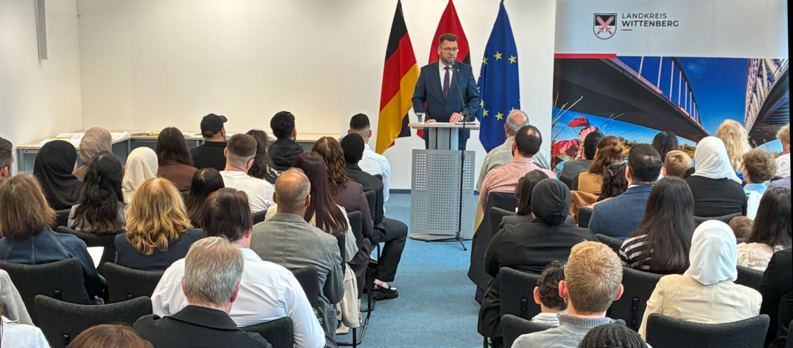 Eine Gruppe Menschen sitzt im Saal, sie sind von hinten zu sehen. in der Mitte steht vorn Landrat Christian Tylsch.