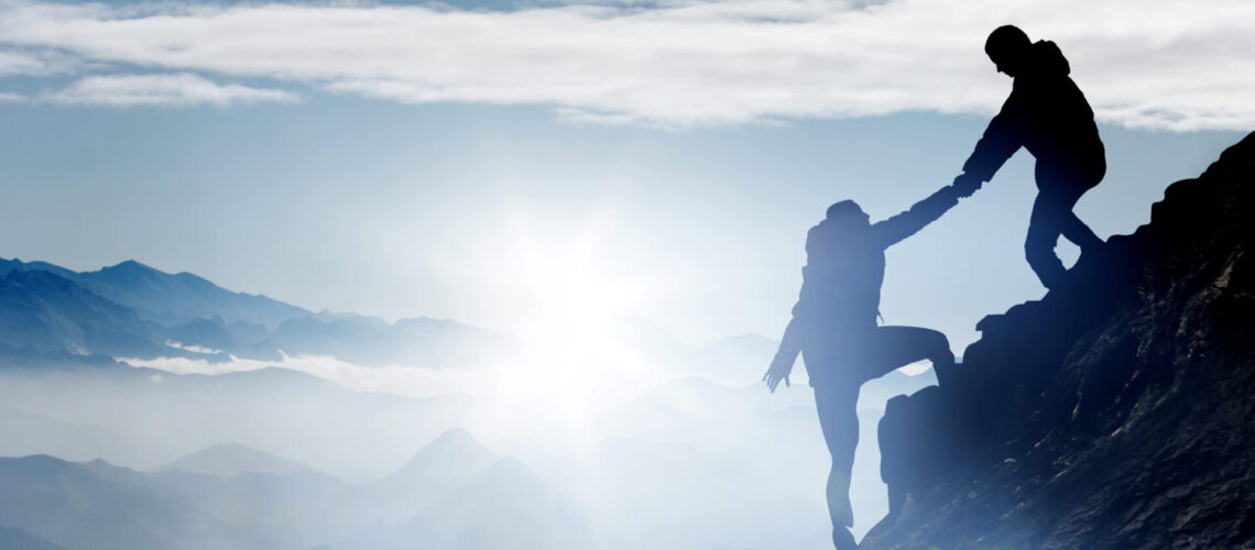 Zwei Menschen beim Bergsteigen und einer davon reicht dem Anderen die Hand und zieht ihn hoch