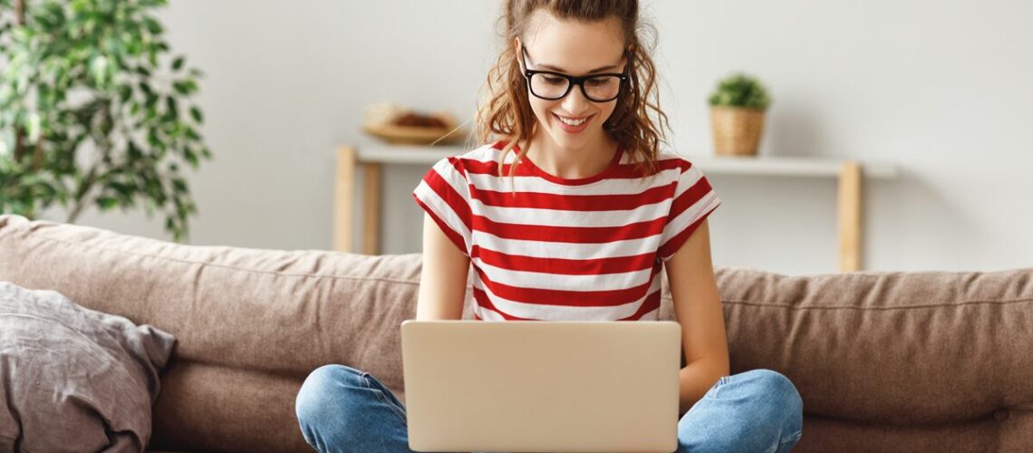 junge Frau sitzt auf einem Sofa im Schneidersitz und schaut auf ihren Laptop