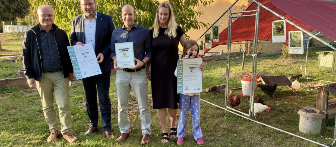 Vier Erwachsene und ein Kind halten Urkunden bzw. eine Auszeichnung in den Händen und lächeln in die Kamera bei der Zertifizierung zur Naturparkschule
