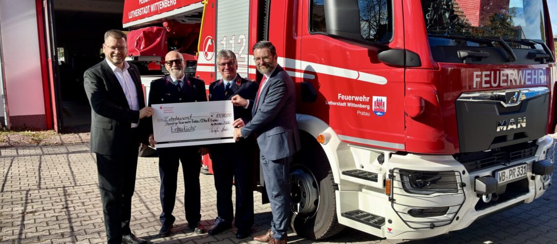 Vier Männer stehen vor einem Feuerwehrauto und halten einen Scheck hoch.