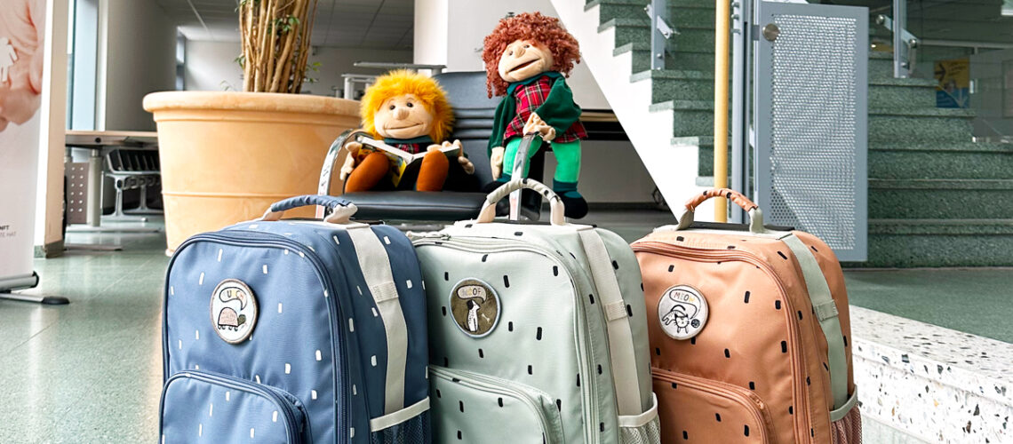 drei Rucksäche in verschiedenen Farben stehen im Vordergrund und im Hintergrund sitzen zwei Puppen auf einem Stuhl